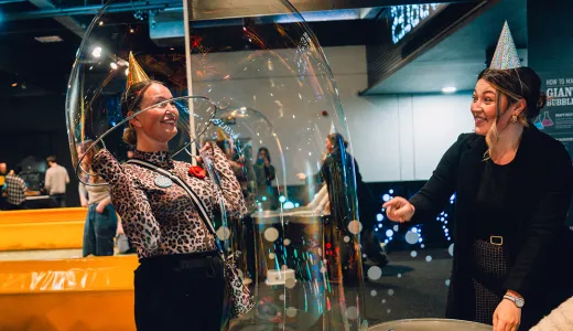 A lady is wearing a gold hat smiling. She is pulling a giant bubble over her head