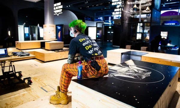A person viewed from behind sat on a wooden box structure in the exhibition space in We The Curious