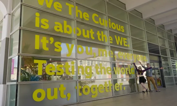 Two people standing relaxed next to a big text on a building that reads We The Curious is about WE. It's you, me everybody. Testing the world out, together.