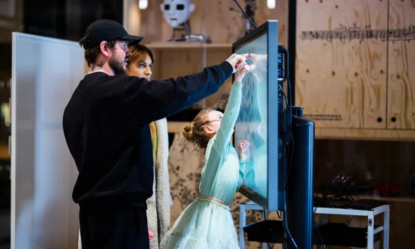 Two adults are helping a child who is reaching up and pointing at something on a screen.