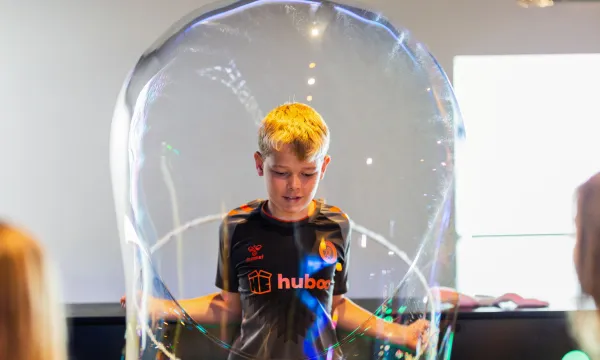An older child is stood in a science centre, pulling a giant bubble over their head.