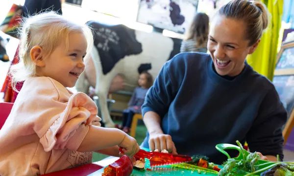 A young child and are smiling and interacting sat at a low table. The child is using a special plastic knife to cut a red pepper.