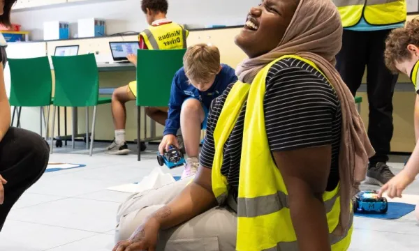 A child is sat on the floor of a classroom wearing a hi-viz jacket and is laughing.