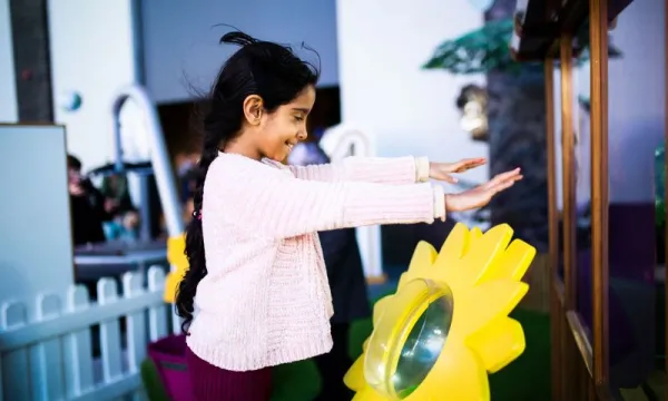 A child is stood in We The Curious science centre in a pink jumper. Stood Infront of a large, yellow, plastic flower and they are holding their hands up over it as it blows wind in their direction.