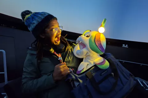 A person is stood in a dark room facing a puppet with their mouth wide open. The puppet is facing back at them with its mouth wide open. The puppet is white, green, and purple and resembles a mushroom-like creature. It has antenna which light up and little white fangs.