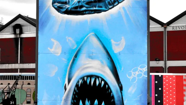 A public artwork showing a shark rising towards the surface towards a large plastic bottle, with red text reading 'Cause'