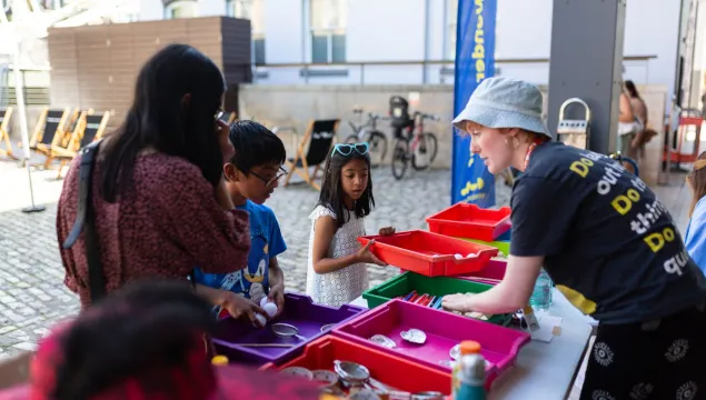 A family group of 3 with a We The Curious staff member, looking at things in a series of coloured trays in an outdoor space