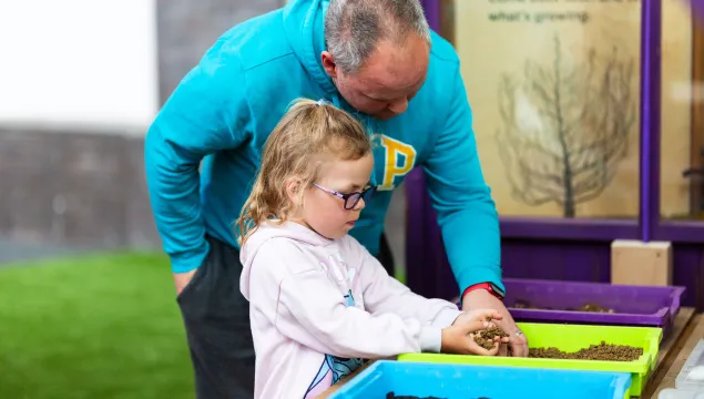 An adult and a child are in a greenhouse playing with soil in plastic containers.