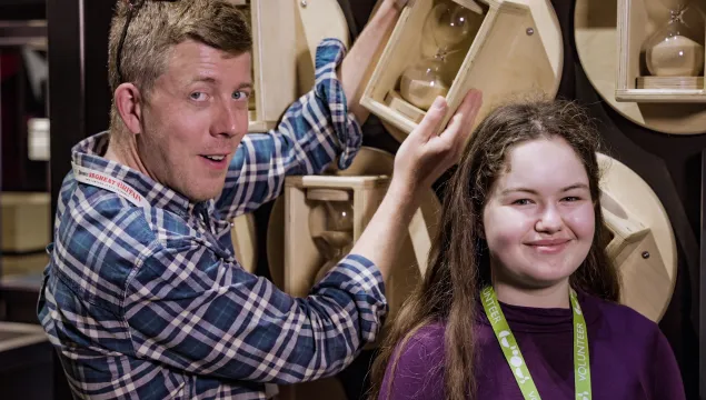 One of the first volunteers of We The Curious smiling and turning an exhibit with a sand clock while one of the latest volunteers is standing in the foreground smiling.  