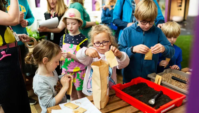 An group of children are in a greenhouse playing with soil in plastic containers and paper bags.