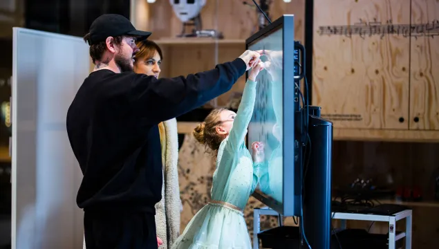 Two adults are helping a child who is reaching up and pointing at something on a screen.