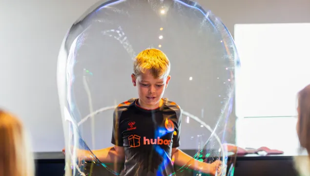 An older child is stood in a science centre, pulling a giant bubble over their head.