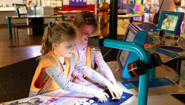 Two children are sat in a science museum in hiz-viz jackets playing with an animation station