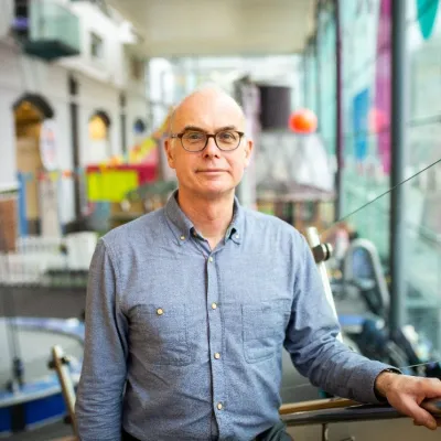 A person smiling with very short white hair and glasses, they are wearing a blue shirt, and are standing in front of a We The Curious exhibit.
