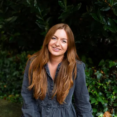 A person smiling with long auburn hair, they are wearing a grey top and are sitting in front of some greenery.