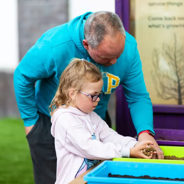 An adult and a child are in a greenhouse playing with soil in plastic containers.