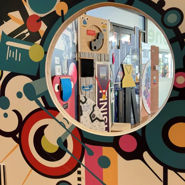 A round window in a colourful wall revealing an art display with geometric shapes, mirrors, and interactive elements in the foyer at We The Curious  